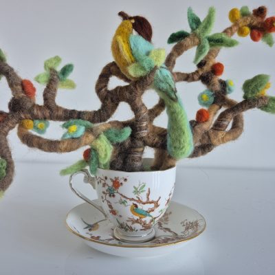 needle felted sculpture of bird in a teacup, brightly coloured felted bird sits in a many branched tree surveying the land from the vintage cup and saucer. brightly coloured leaves and flowers adorn the tree of this needle felted sculpture in a teacup.